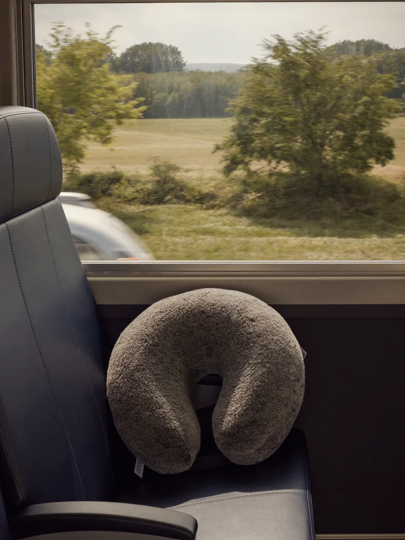 Nackenkissen mit grauem Strickbezug auf einem Zugsitz vor Fensterlandschaft.